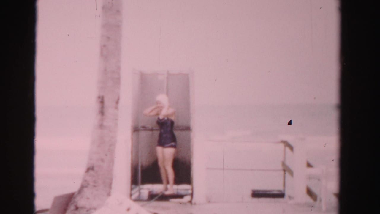 Woman in a dark swimsuit and swim cap stands in a poolside doorway near a palm tree.