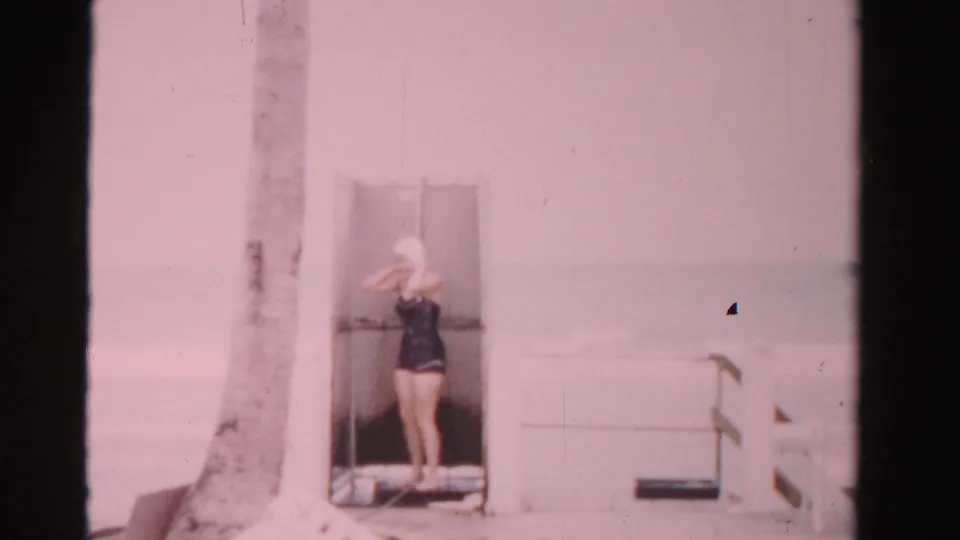 In 1955 Dade County, Florida, a woman in a dark one-piece swimsuit and swim cap stands by a changing-room doorway near a palm tree.