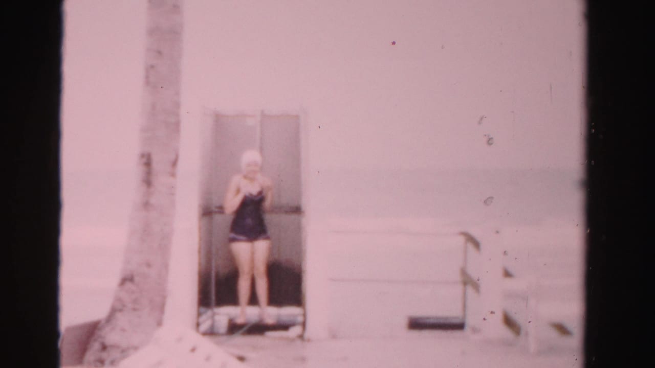 The woman remains at the poolside doorway in Dade County, Florida, adjusting near her swim cap against a pale wall and palm trunk.