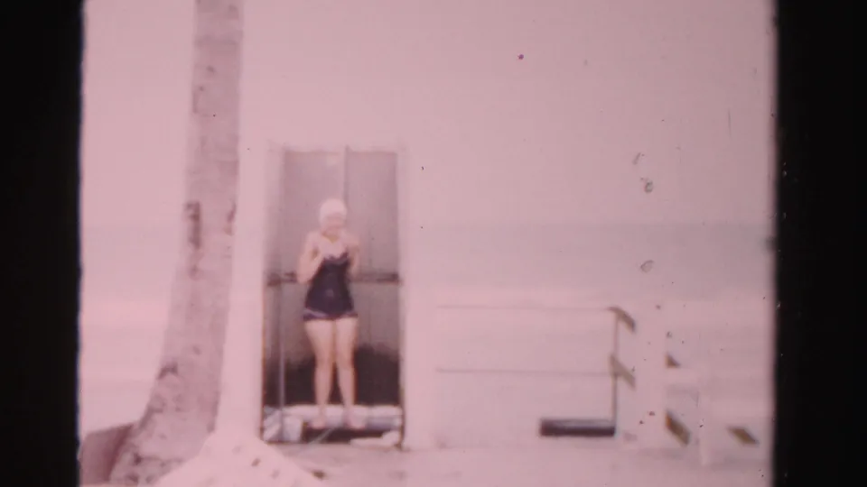 The woman remains at the poolside doorway in Dade County, Florida, adjusting near her swim cap against a pale wall and palm trunk.
