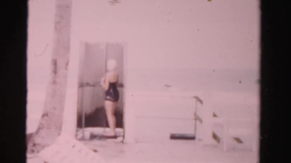 The swimmer turns partly away in the changing-room doorway, with the palm trunk and simple poolside structure visible around her.