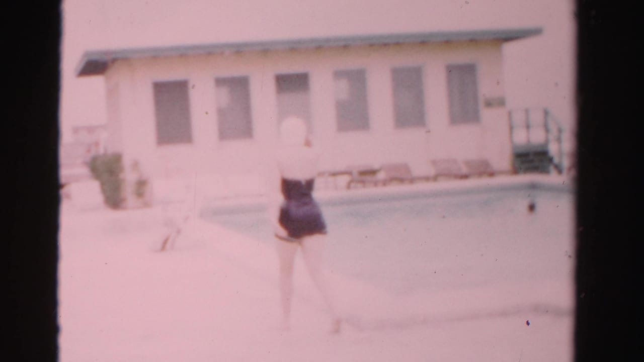 Woman in a dark swimsuit and swim cap walks along a bright pool deck beside a pale building.