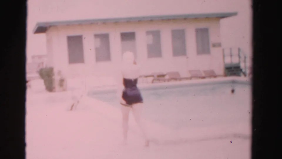 In 1955 Dade County, Florida, a woman in a one-piece swimsuit and swim cap approaches a swimming pool near a light-colored building.