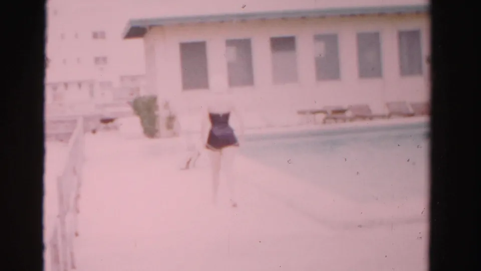 The woman moves farther along the Dade County pool deck, filmed in soft, pink-tinted 8mm with a pale building behind her.