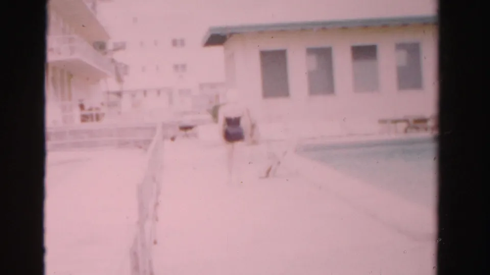 At the end of the clip, the woman remains near the pool edge, framed by the light building and deck furniture in 1955 Florida footage.