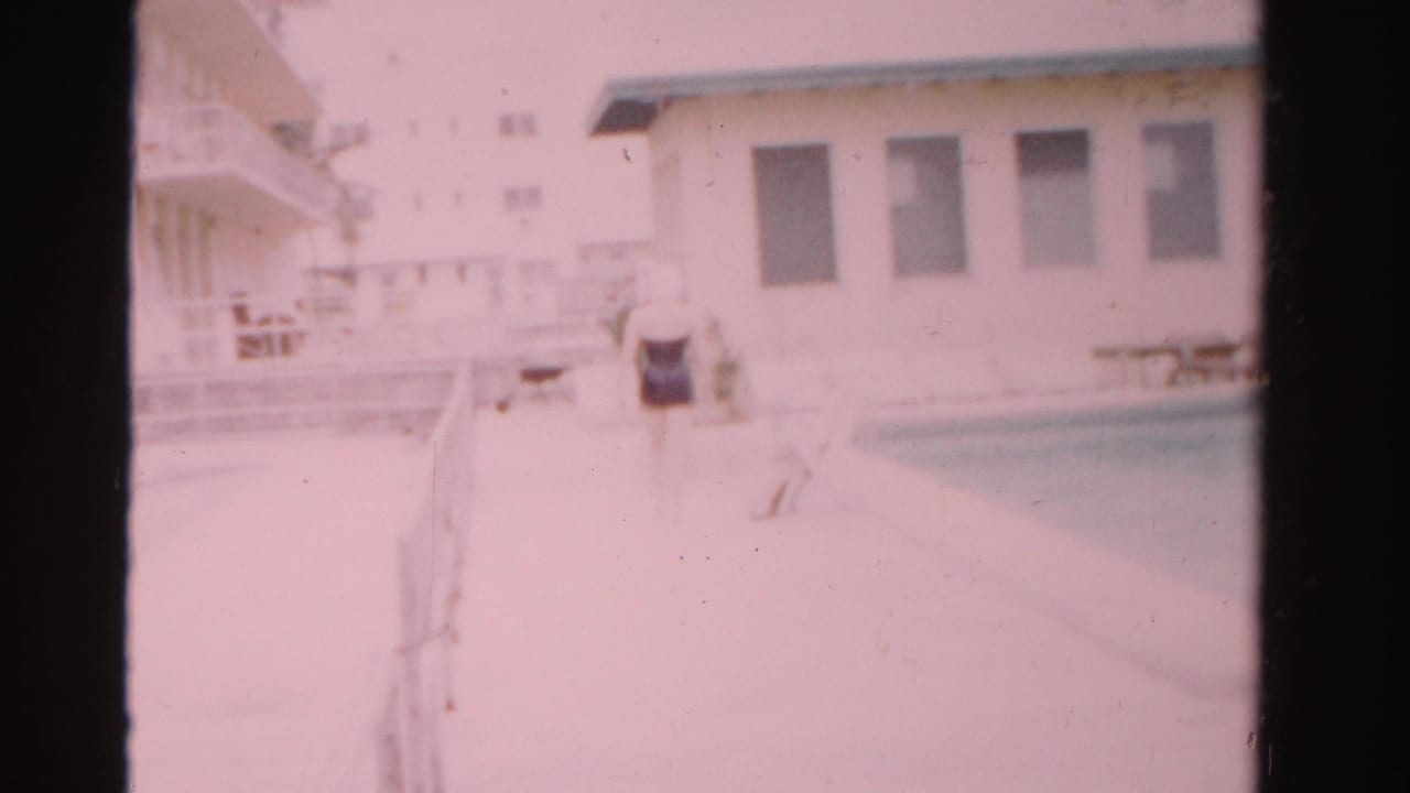 Woman in a dark swimsuit walking along a bright pool deck beside a white building.
