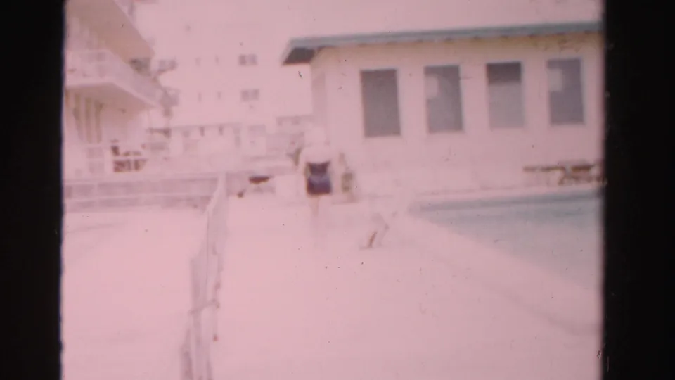 1955 Dade County 8mm frame showing a woman in a dark swimsuit on a sunlit pool deck near a white building and railings.