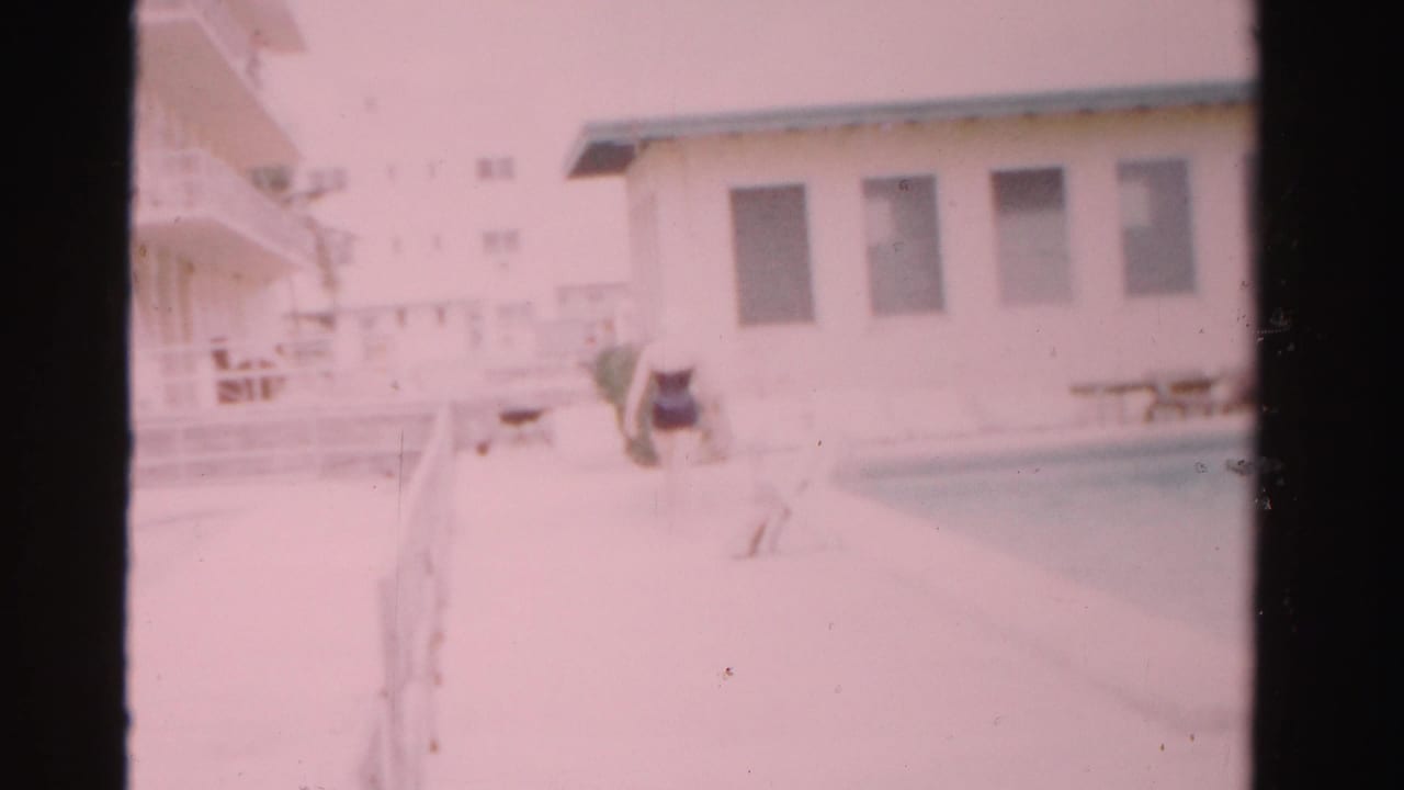 Woman approaching the edge of a swimming pool near a white building and outdoor deck.