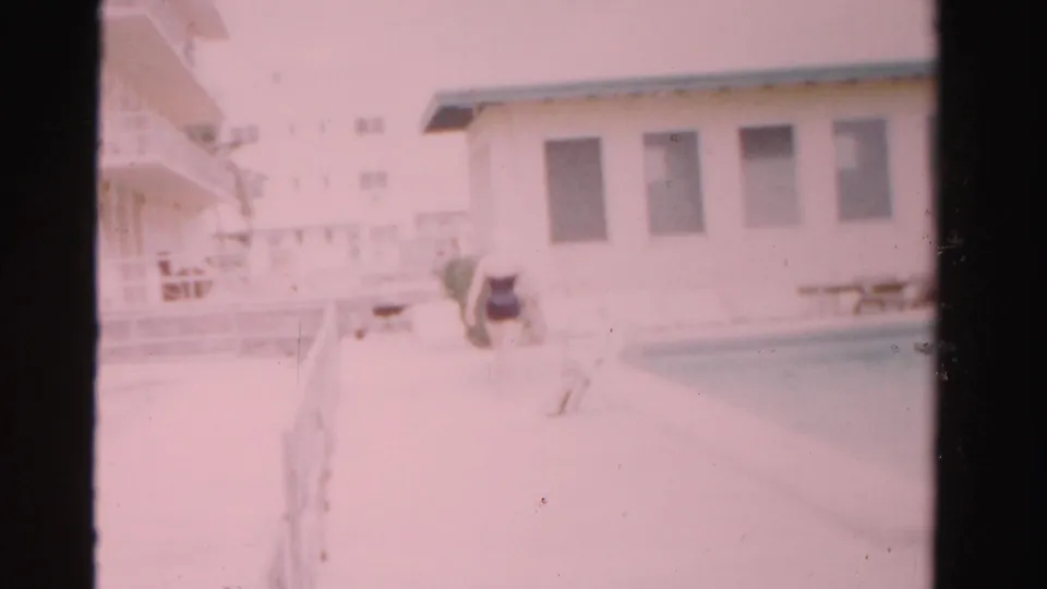 1955 Dade County poolside frame with a woman nearing the water, a white building, windows, railings, and deck furniture visible.