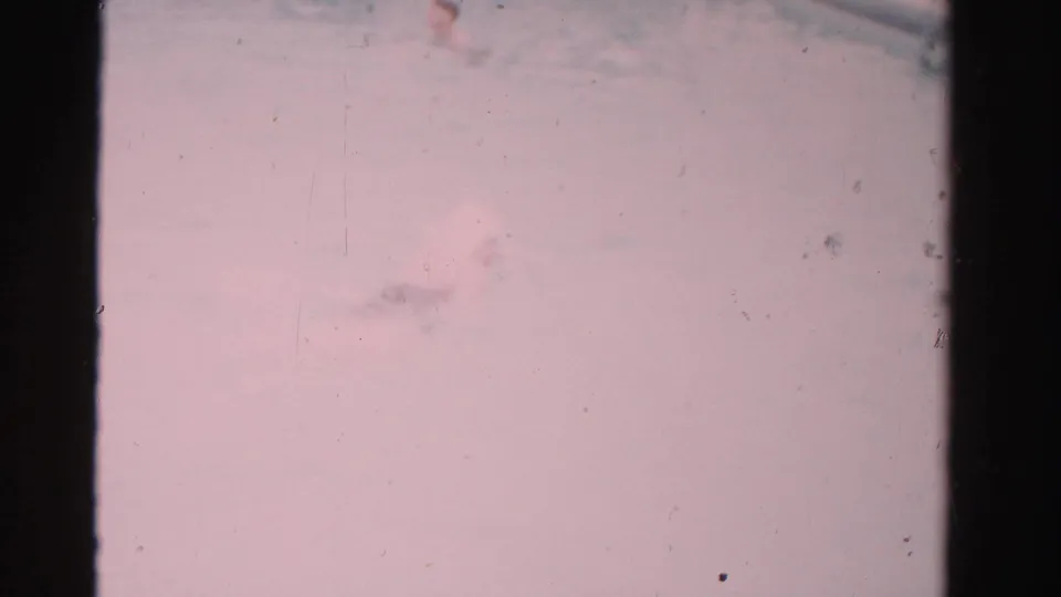 1955 Dade County home-movie frame showing a pale pink poolside or water surface area, dark gate edges, dust, and faded 8mm color.