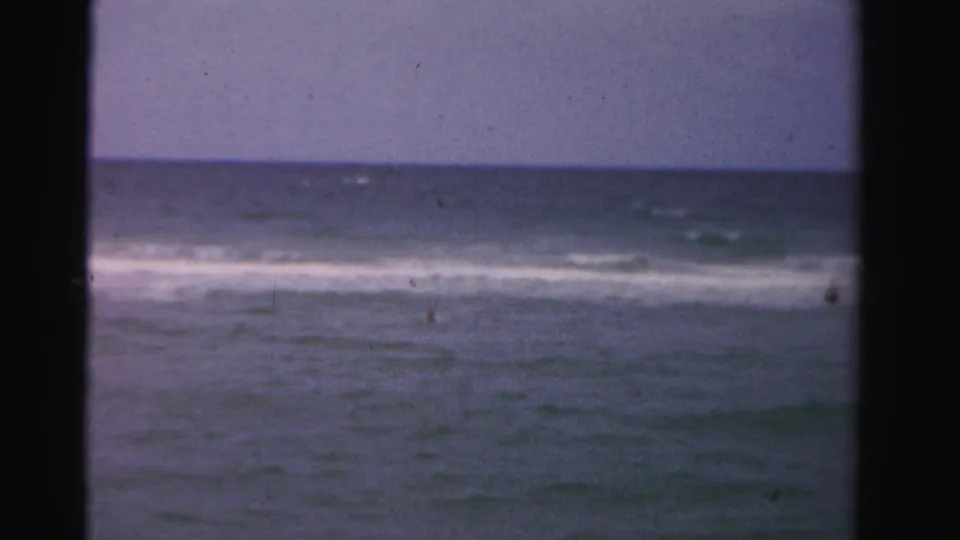 Wide 1955 Dade County home-movie view of ocean water, white surf, and a few distant swimmers under faded blue color.