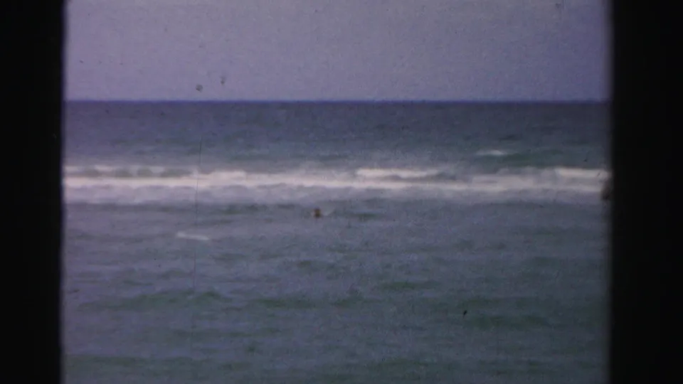 1955 Dade County ocean frame with faded blue water, a white surf band, and a small distant swimmer visible near center right.