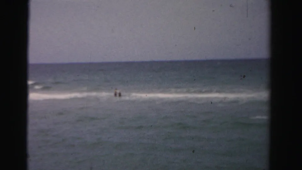 1955 Dade County beach footage with two distant figures in shallow ocean water near small breaking waves.