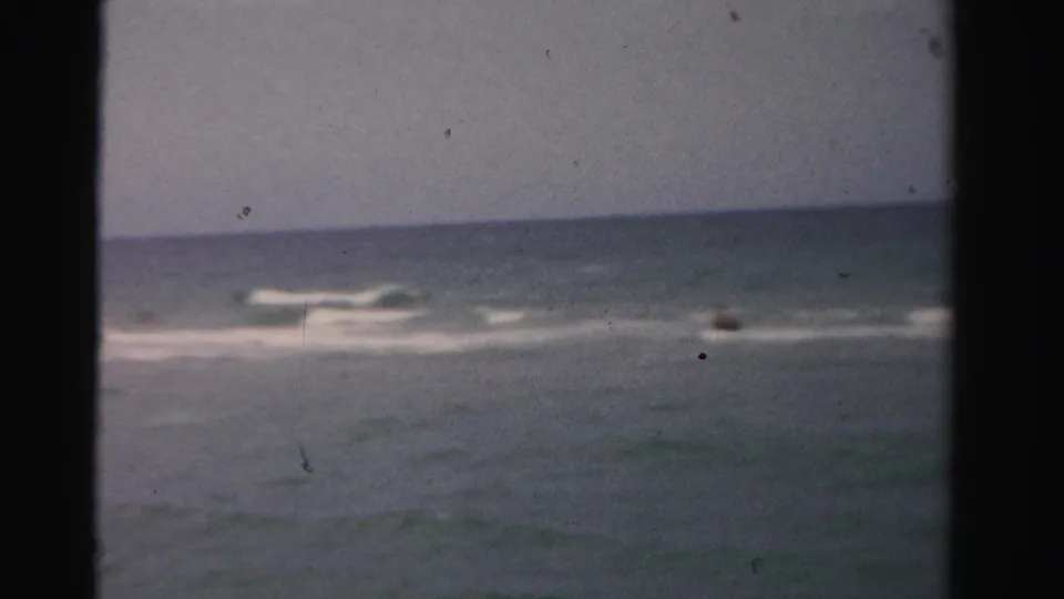 Faded 8mm Dade County ocean view with low surf and a distant dark shape that may be a swimmer or object in the water.