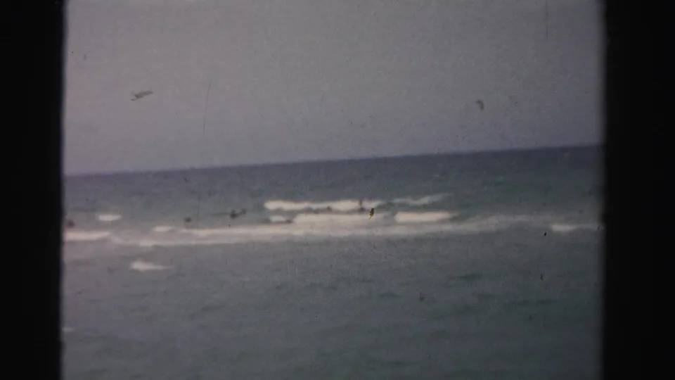 1955 Dade County 8mm frame showing a distant swimmer in calm ocean water under a muted sky.