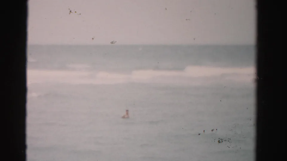 A distant swimmer appears in the ocean off Dade County, Florida, with soft waves, overcast light, and visible 8mm film borders.