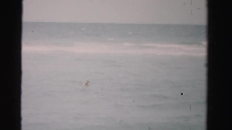 1955 8mm footage shows a distant swimmer moving through muted blue-gray ocean water near Dade County, Florida.