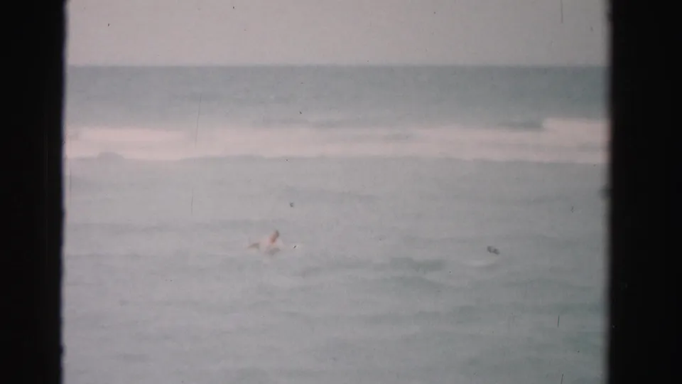 A distant swimmer moves through small ocean waves in this 1955 Dade County home-movie frame with grain and soft exposure.