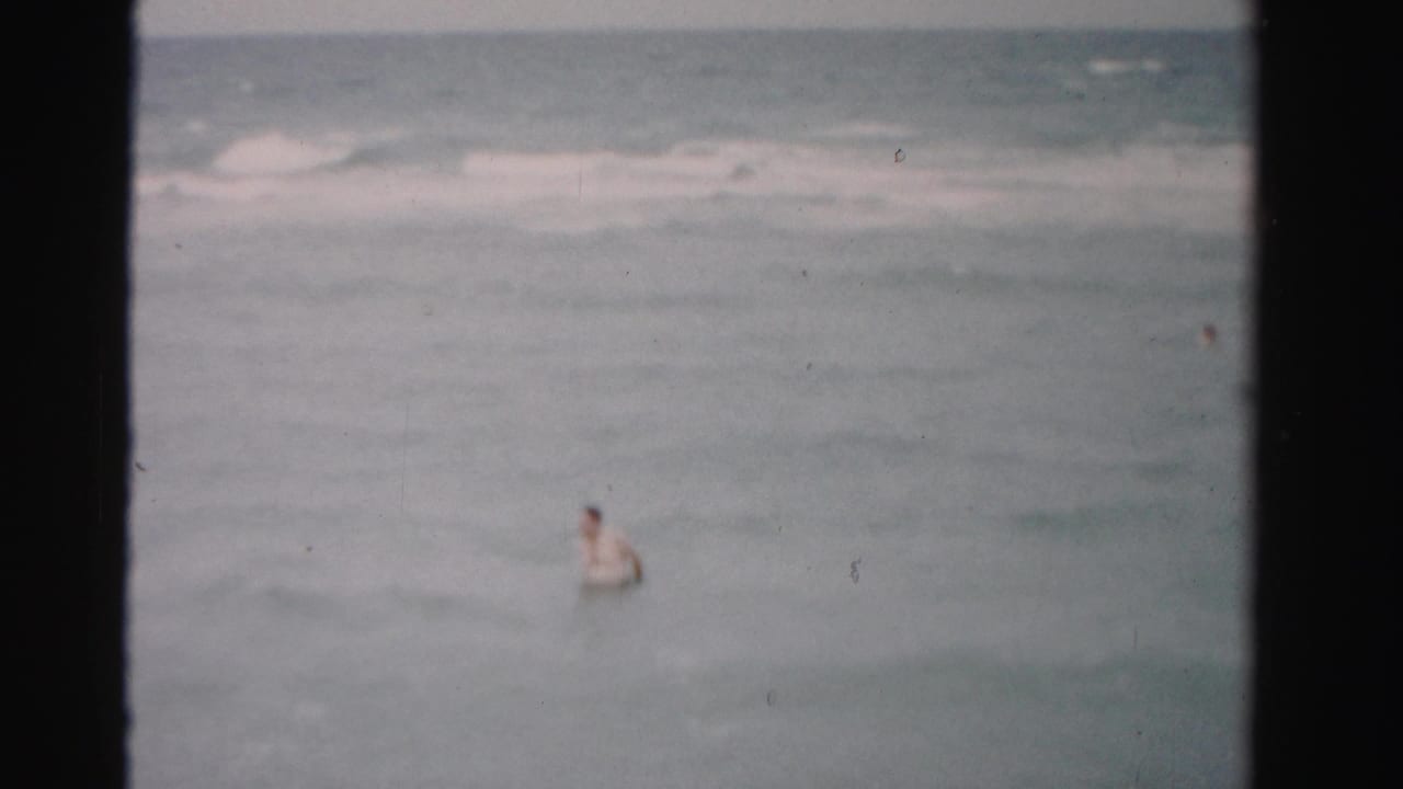 A man stands or floats in pale ocean surf, framed by dark 8mm film borders.