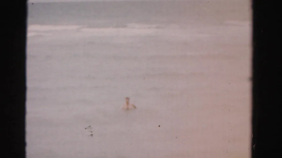 A swimmer appears offshore in a faded 1955 Florida beach home-movie frame with waves behind him.