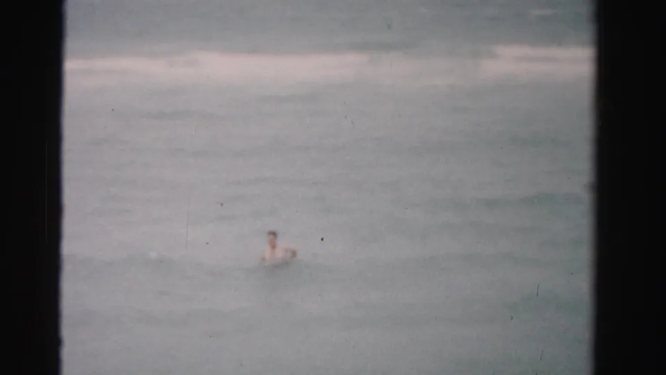 The home-movie frame shows a swimmer low in the shot with broad ocean surf and 8mm gate edges.