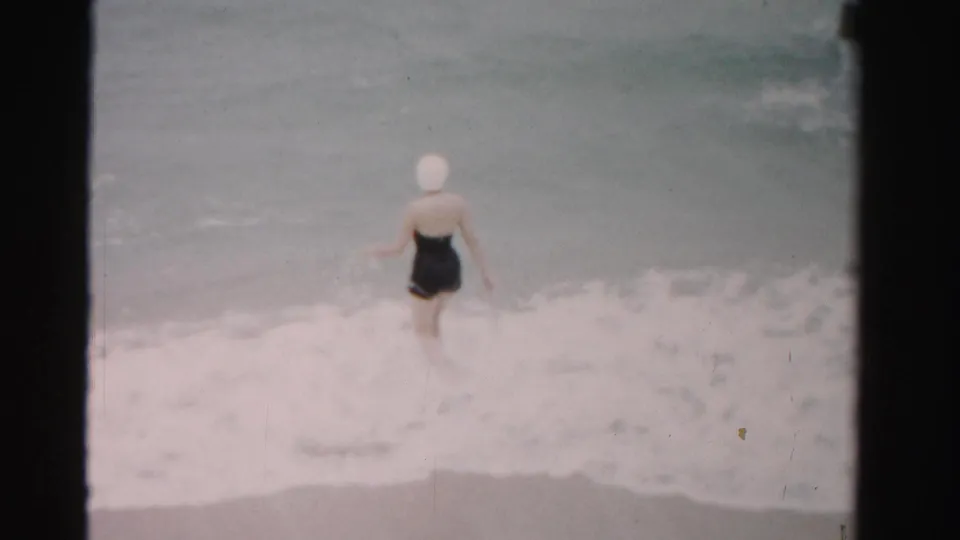 In 1955 Dade County beach footage, a woman in a dark swimsuit and white cap steps into the ocean surf.