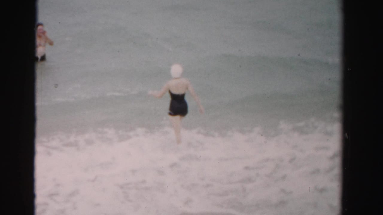 Woman in a black swimsuit wades into shallow surf while a man stands farther out in the water.