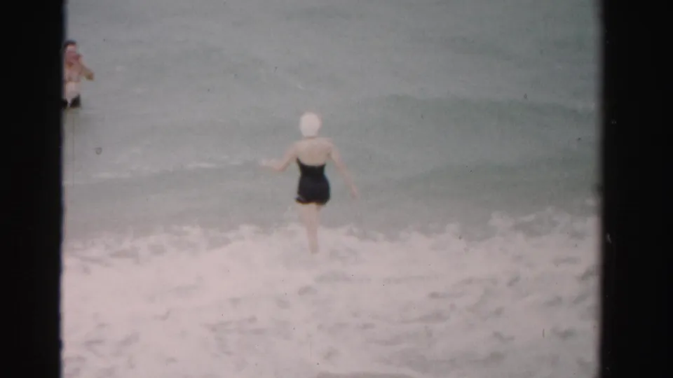 1955 Dade County home-movie frame showing a woman in a black swimsuit and white swim cap entering the surf as a man waits offshore.