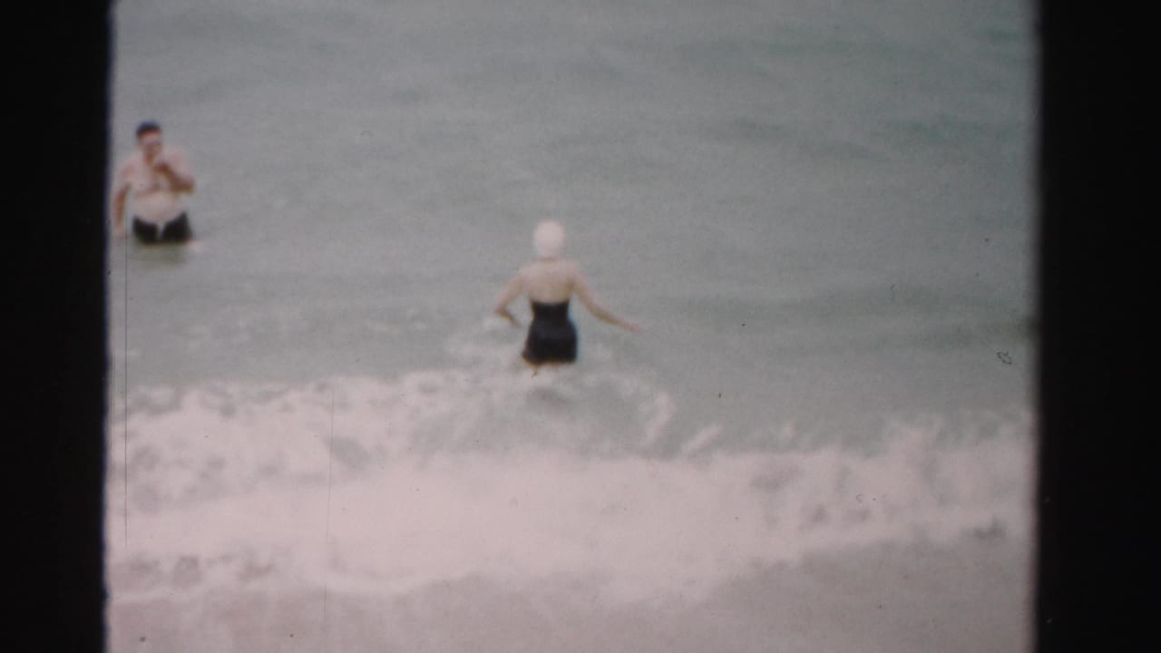 Two adults in swimwear stand in shallow ocean water as a wave breaks near the shore.