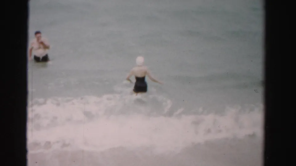 1955 Dade County beach frame with a woman in a white swim cap and a man in dark trunks standing apart in gentle surf.