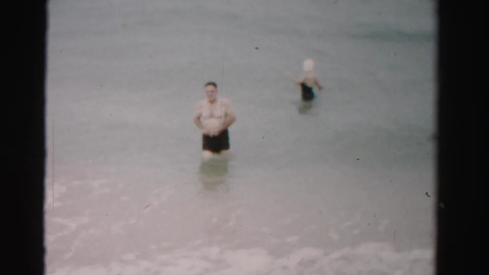 1955 Dade County home-movie frame of a man in dark swim trunks and a woman farther out in shallow ocean water.