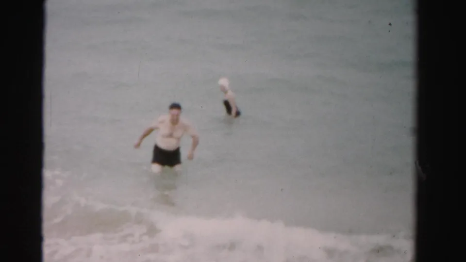 1955 Dade County beach frame with a man moving through shallow surf and a woman standing farther out in the water.