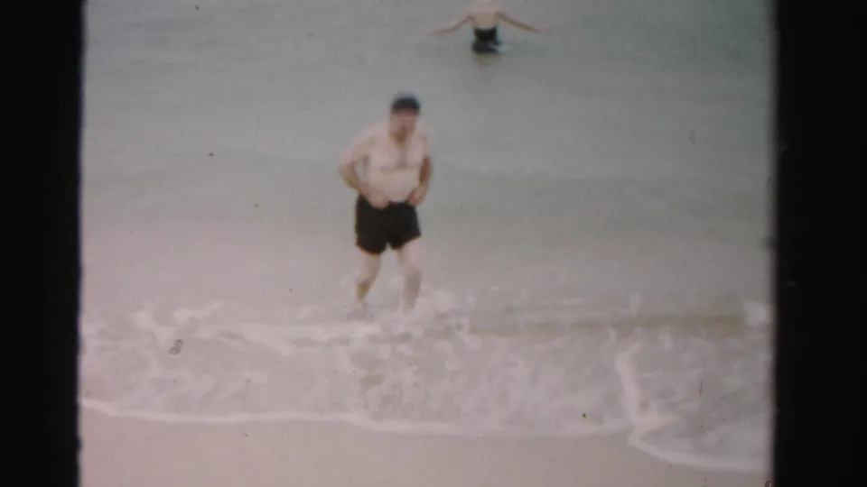 1955 Dade County home-movie frame of a man stepping through foamy shoreline water with a woman partly visible offshore.