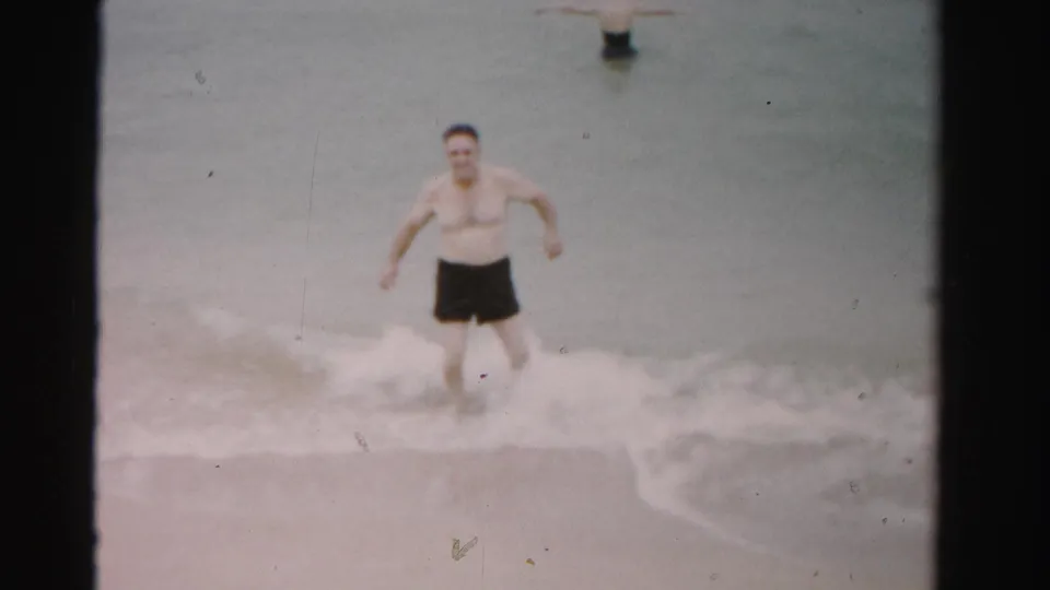 Adult man in dark swim trunks moves through low ocean waves in 1955 Dade County, Florida home-movie footage.