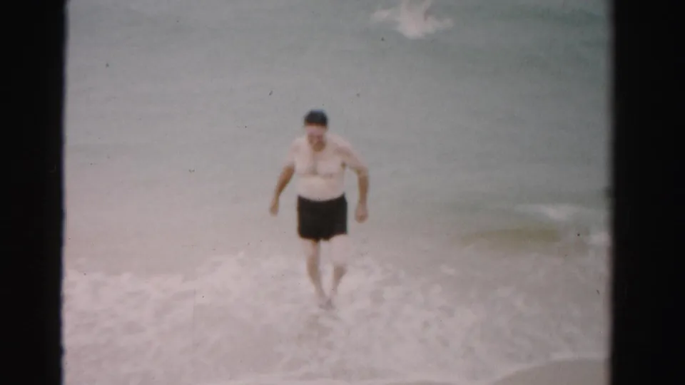 Man in dark swim trunks steps through foamy beach water in the Dade County, Florida surf.