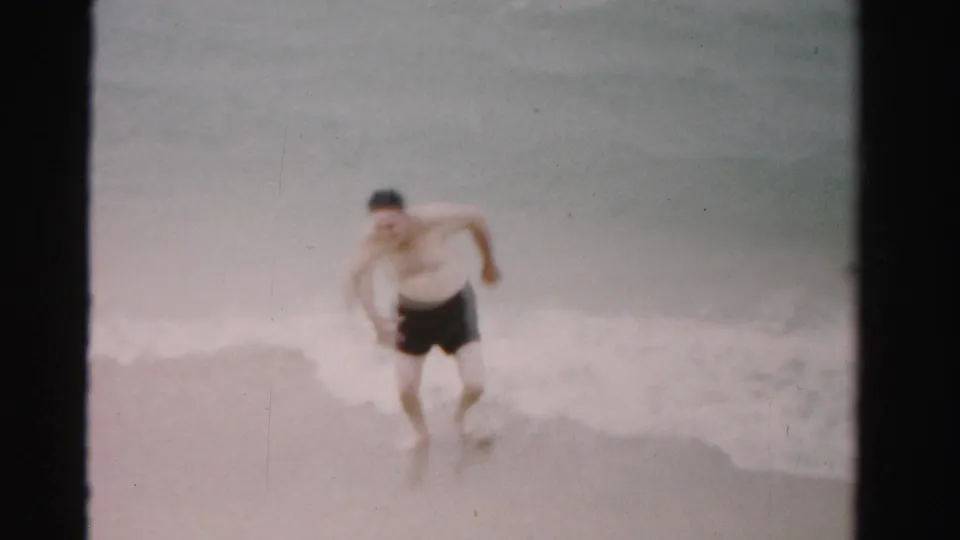 Adult beachgoer in dark trunks leans into motion at the surf line in 1955 Florida home-movie footage.