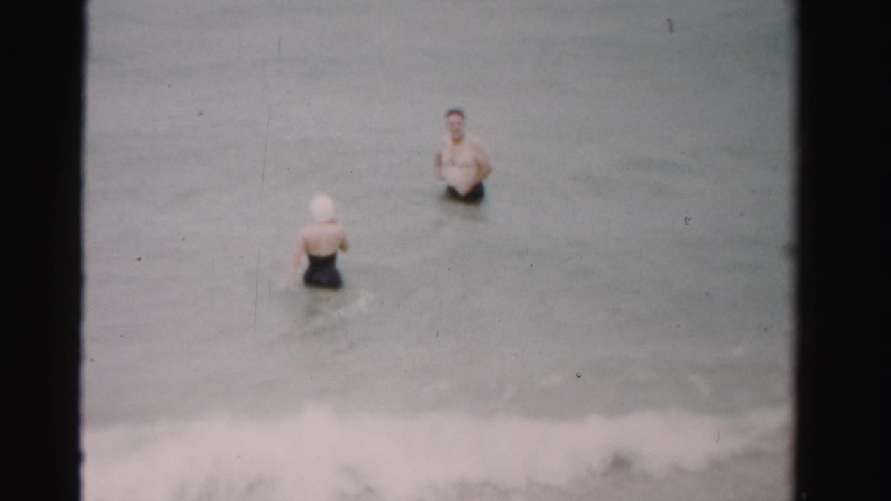1955 Dade County home-movie frame showing a woman in a white swim cap and a man in swim trunks standing in shallow surf.