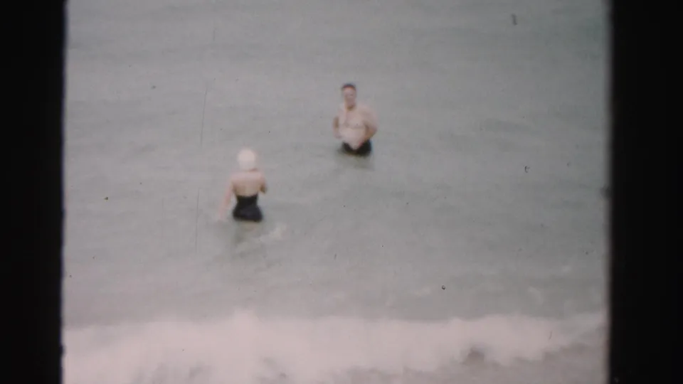 1955 Dade County home-movie frame showing a woman in a white swim cap and a man in swim trunks standing in shallow surf.