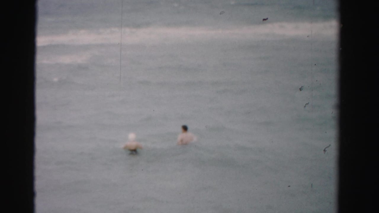 1955 Florida beach frame with two adults wading farther into the ocean, seen small and soft-focused from shore.