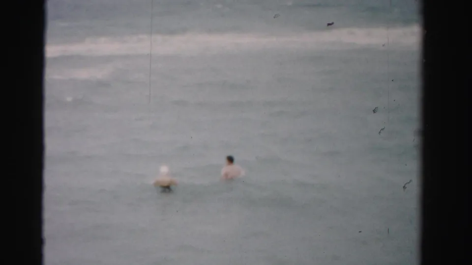 1955 Florida beach frame with two adults wading farther into the ocean, seen small and soft-focused from shore.