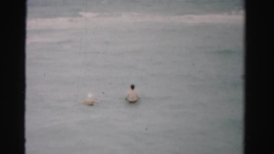 1955 Dade County beach footage frame showing two adults in swimwear moving through the surf under soft 8mm color.