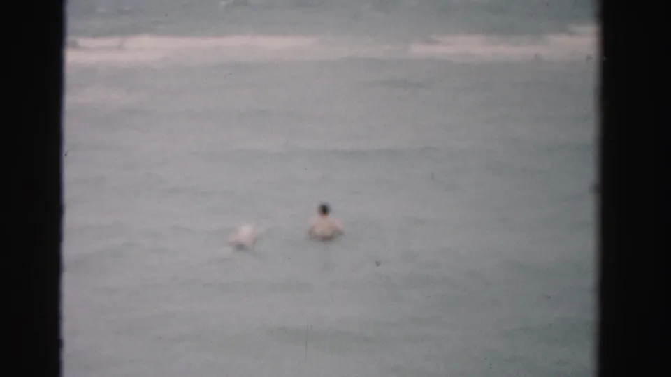 Soft-focused 1955 Florida home-movie frame of two adults partly submerged while wading in open surf.