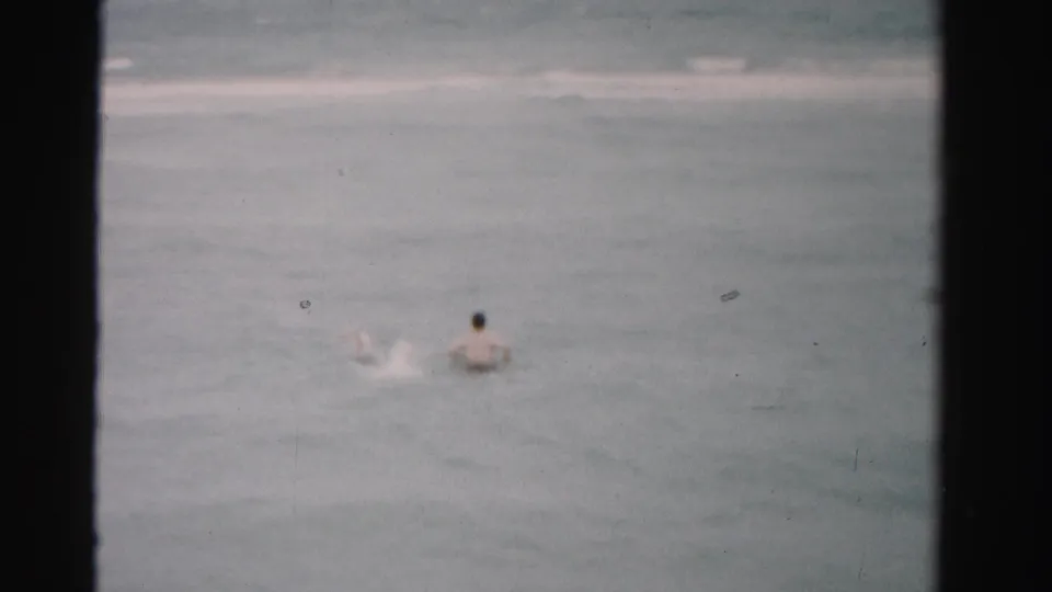 A distant swimmer appears in muted 1955 Dade County, Florida surf, framed by 8mm film borders and light surface wear.