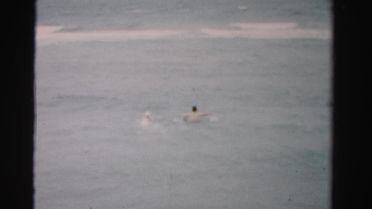 1955 Dade County home-movie frame showing two distant swimmers in gray-green ocean water under cloudy light.