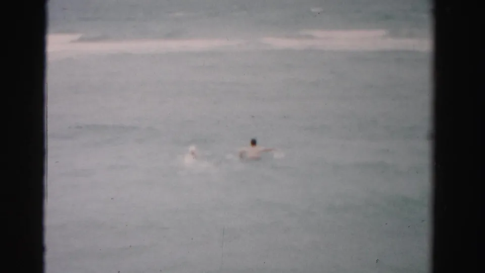 1955 Dade County home-movie frame showing two distant swimmers in gray-green ocean water under cloudy light.