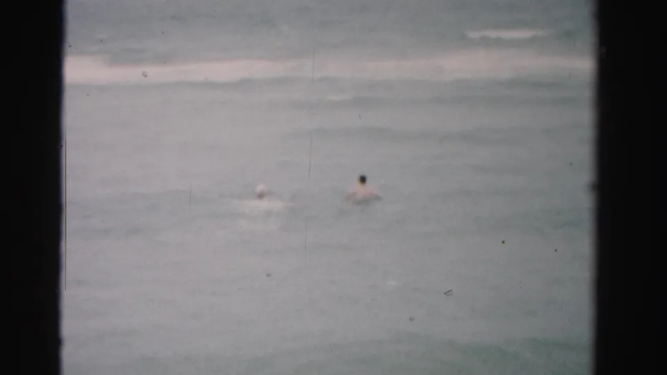 Distant swimmers in 1955 Dade County ocean footage, seen through faded 8mm color with dust and gate-edge borders.