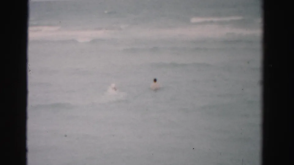 1955 Dade County home-movie frame showing two distant people swimming in muted ocean water under a gray sky.