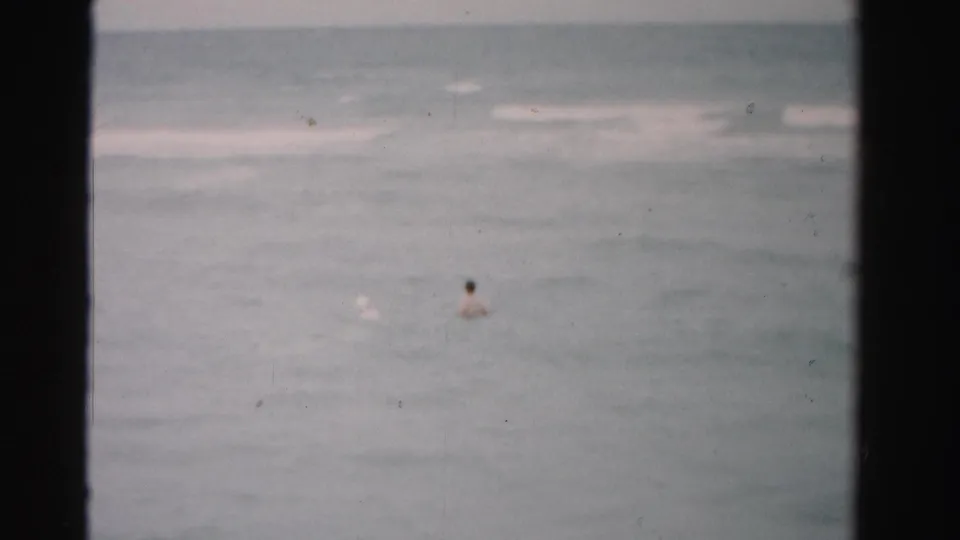 Dade County 1955 home-movie still showing distant swimmers in the ocean, with soft focus and visible film texture.