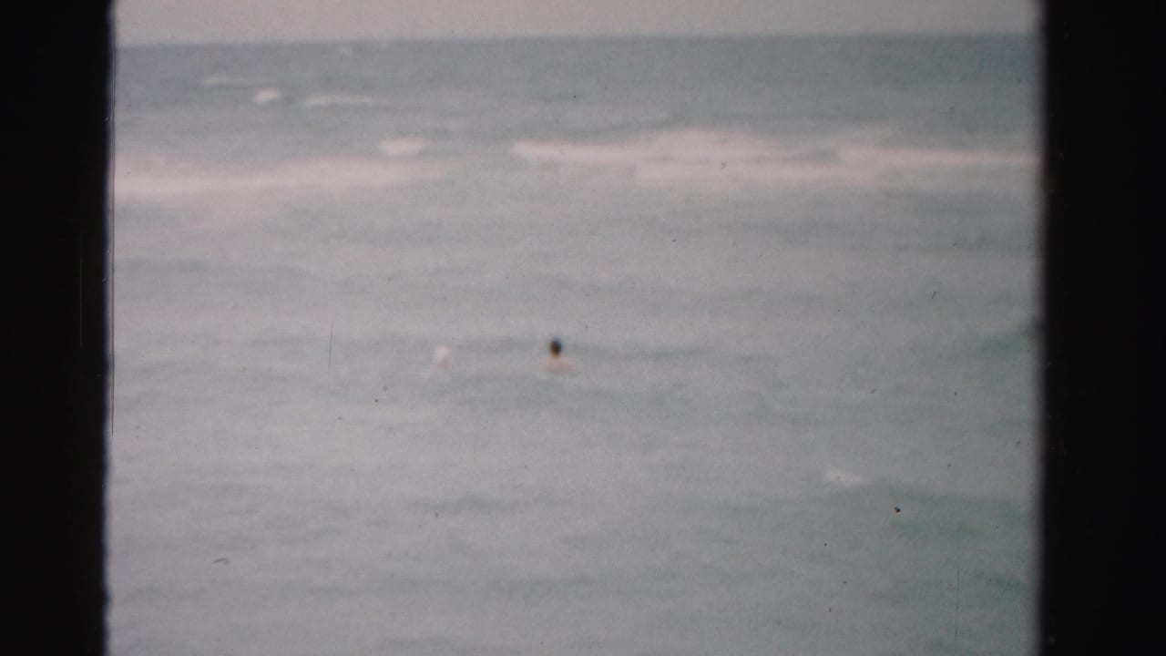 Final 1955 Florida 8mm frame with two distant swimmers in gray-green sea water and low surf near the horizon.