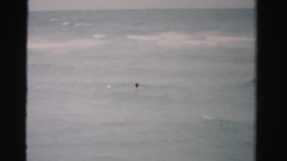 Final 1955 Florida 8mm frame with two distant swimmers in gray-green sea water and low surf near the horizon.